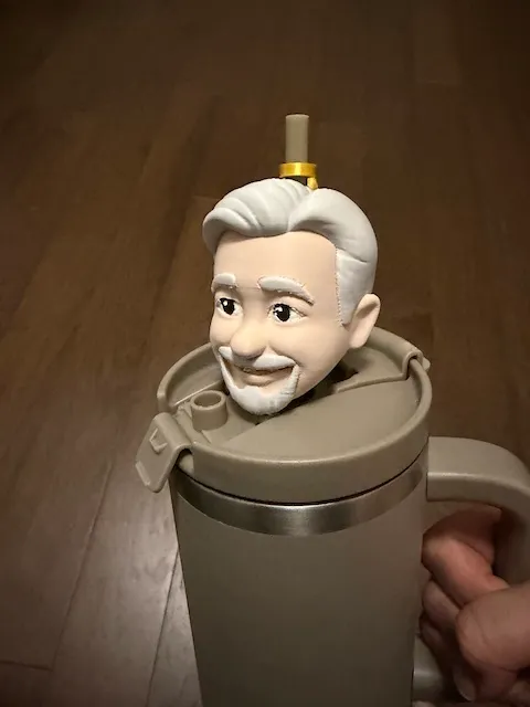 A 3D-printed bust of a smiling white-haired man with a mustache and goatee sits in the lid opening of a Stanley-style tumbler, with the drinking straw emerging from the top of its head.